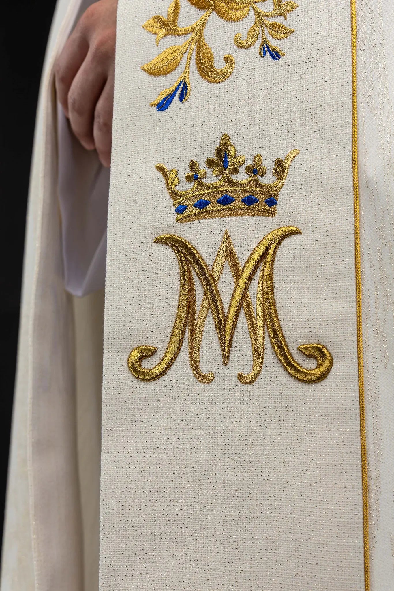 Ecru embroidered cope with Marian symbol, featuring gold and blue floral embroidery, a crowned 'M' monogram, and an ornate gold clasp with chain.
