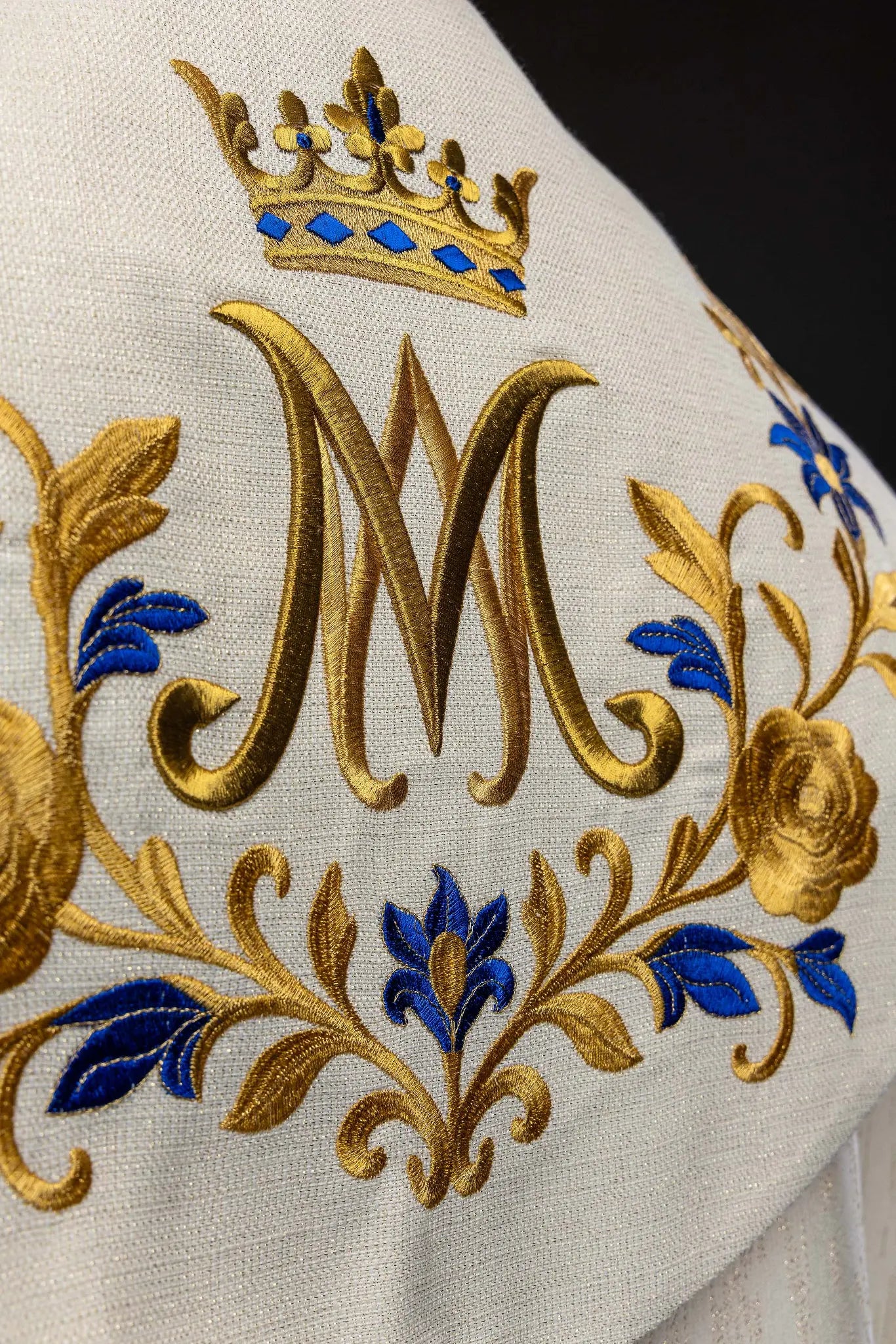 Ecru embroidered cope with Marian symbol, featuring gold and blue floral embroidery, a crowned 'M' monogram, and an ornate gold clasp with chain.